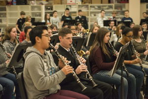 Eine Gruppe von Menschen, die Klarinetten in einem Klassenzimmer spielen, einige sitzen und andere stehen, mit Notenpulten und Regalen mit Kisten im Hintergrund.