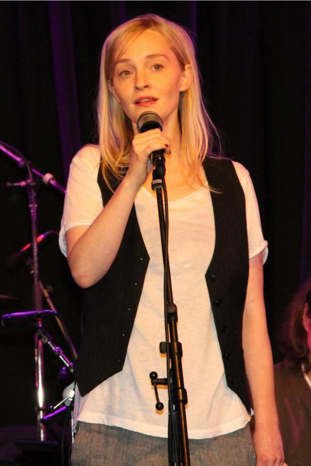 Frau auf einer Bühne mit einem Mikrofon stehend, die eine weiße T-Shirt und eine schwarze Jacke trägt, mit einer anderen Person rechts von ihr und Musikinstrumenten im Hintergrund.