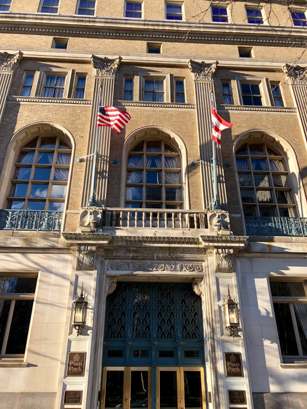 Großes Gebäude mit Glasfenstern, -türen, -säulen, -geländern, -leuchten; mehrere Flaggen an der Vorderseite; Texttafeln und das Plaza Hotel-Schild; vor einem Himmel-Hintergrund.