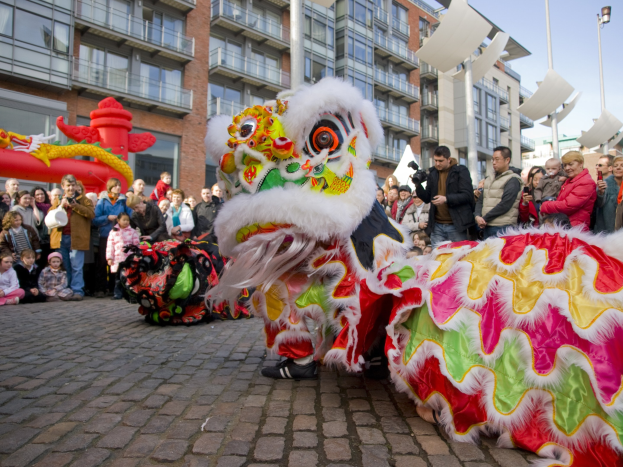 Ein lebendiges chinesisches Neujahrsfest in Amsterdam mit einer Löwen-Tanzdarbietung vor einer Zuschauermenge, einige mit Kameras, vor einer Kulisse aus Gebäuden, Laternenmasten und einem klaren blauen Himmel.