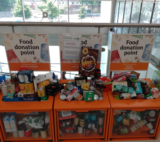 Orange Schübe mit Lebensmittelspenden gefüllt, darunter Flaschen, Dosen und Kisten, mit informativen Schildern im Hintergrund, vor dem Fenster eine Aussicht auf Fahrzeuge, Bäume, Gebäude und Himmel.