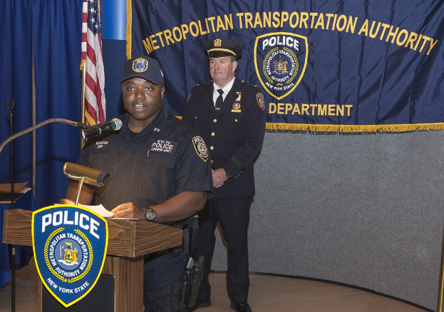 Zwei Polizeibeamte auf einem Podium mit einem Banner, einer Fahne und einer Tafel mit Symbolen und Text, einer Beamter spricht in ein Mikrofon und hält Papiere.