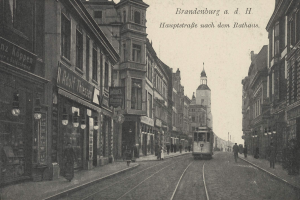 Schwarz-weiß-Foto einer Straßenkreuzung in Brandenburg, Deutschland mit einer Tram, Passanten, Gebäuden, Schildern, Straßenlaternen und einem bewölkten Himmel.
