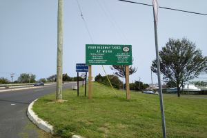 Eine Autobahn mit Fahrzeugen, Gras, Pfählen, Schildern, Bäumen und Strommasten mit Drähten, auf der eine Tafel mit der Aufschrift "Ihre Autobahnsteuer" zu sehen ist.