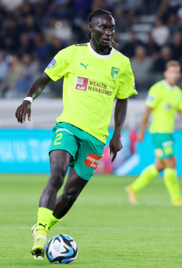 Ein Fußballspieler in einem gelben T-Shirt und grünen Shorts ist im Einsatz auf dem Feld, mit einer blauen Tafel und unscharfen Figuren im Hintergrund.