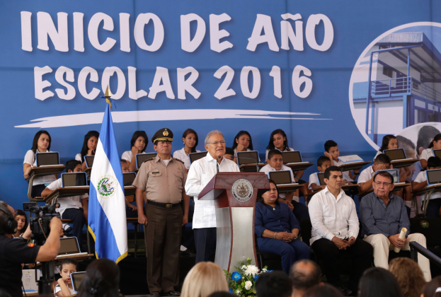 Ein Mann steht an einem Rednerpult und spricht zu einer Menge, mit einer Fahne, einem Banner und einer Person mit einer Videokamera im Hintergrund, wahrscheinlich der Präsident von El Salvador bei der Inicio de Año Escolar 2016.