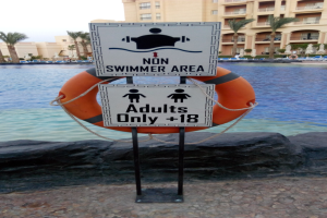 Ein Schwimmbad mit einem "Nur für Nichtschwimmer Bereich Erwachsene"-Schild im Vordergrund, umgeben von Bäumen, Gebäuden und einem klaren blauen Himmel, mit einem Rettungsring in der Mitte des Pools.