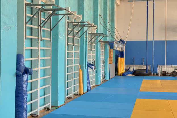 Turnhalle mit blauen und gelben Matten, Leitern, Stangen, Seilen und anderem Equipment an einer Wand und Decke.