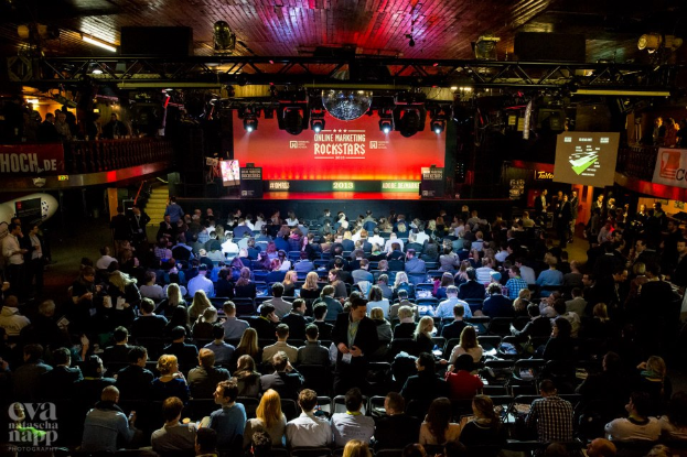 Eine große Menschenmenge sitzt auf Stühlen, die einer Bühne mit Bildschirmen und Plakaten zugewandt sind, beleuchtet von Scheinwerfern und umgeben von Geländern, mit einem Wasserzeichen in der unteren linken Ecke, das eine Atmosphäre der Aufregung und Vorfreude für ein Online-Marketing-Rockstars-Event schafft.
