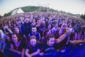 Menschenmenge auf einem Musikfestival mit erhobenen Händen vor einem Geländer, Bäume und Hütten im Hintergrund unter einem klaren blauen Himmel.