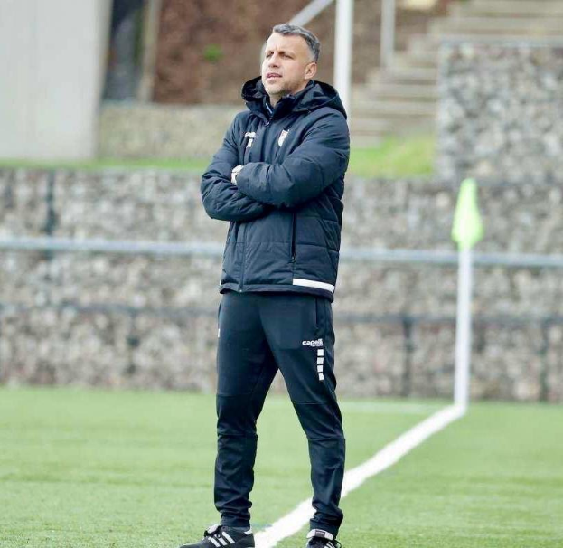 Ein Fußballtrainer steht auf einem Feld und hat die Arme verschränkt, trägt eine schwarze Jacke und Hose.
