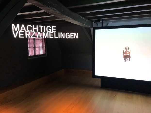 Installationsansicht der Ausstellung 'Machtige Sammlungen' im Museum of Modern Art in Berlin, mit einem großen Bildschirm mit Wandtext, einem Fenster links und sichtbarer Decke.
