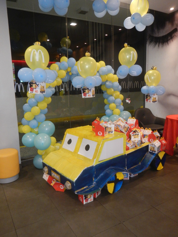 Eine McDonald's-Geburtstagsparty-Szene mit einem gelben und blauen Lastwagen, der mit Ballons und anderen Dekorationen geschmückt ist, einem rot gedeckten Tisch mit Gegenständen, Hockern, einer Glaswand und Deckenlampen.