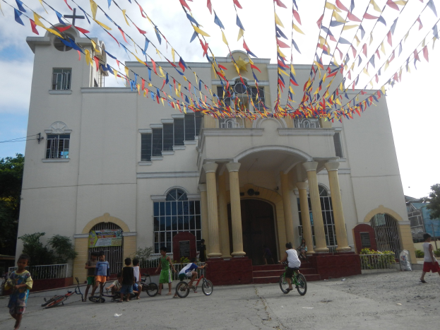 Eine Gruppe von Menschen, die auf Fahrrädern vor einer weißen Kirche mit architektonischen Details fahren, umgeben von Grün, einem Banner, Bäumen und einem bewölkten Himmel, mit bunten Fahnen oben.