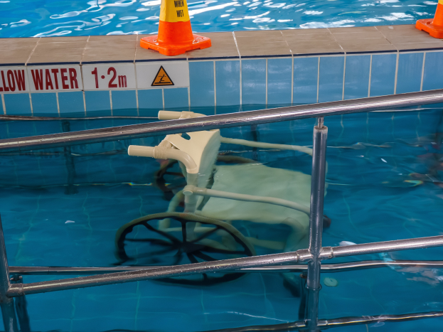 Schwimmbad mit einem Stuhl in der Mitte, umgeben von Sicherheitstonnen und einem Geländer, mit einem Schild an der Wand, das verfügbare Schwimmausrüstung anzeigt.