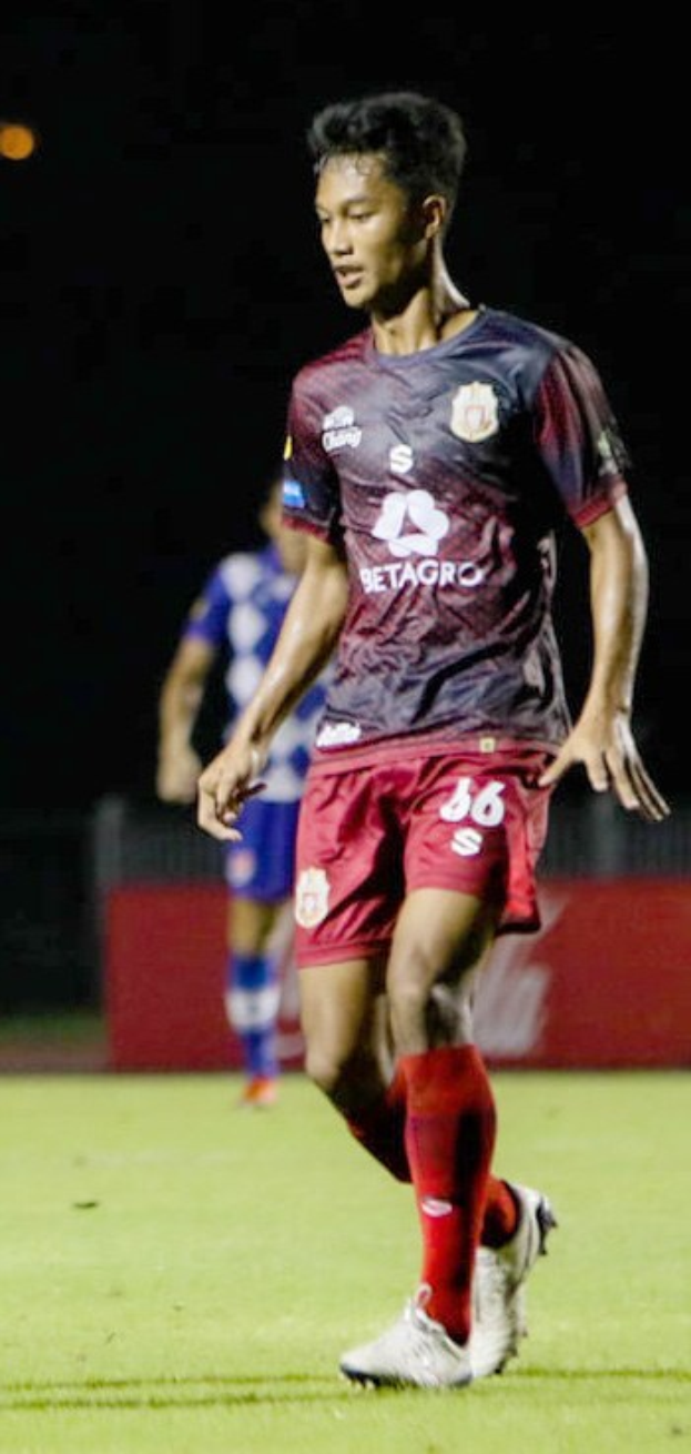 Ein Mann in einem roten Hemd und kurzen Hosen, der auf einem Feld Fußball spielt, mit einer anderen Person im Hintergrund und einer Tafel mit Text.