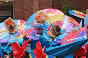 Eine Gruppe von Kindern in bunten Kostümen fährt auf einem Paradewagen, mit Schirmen, vor einem Gebäude, Pflanzen und Lichtern im Hintergrund.