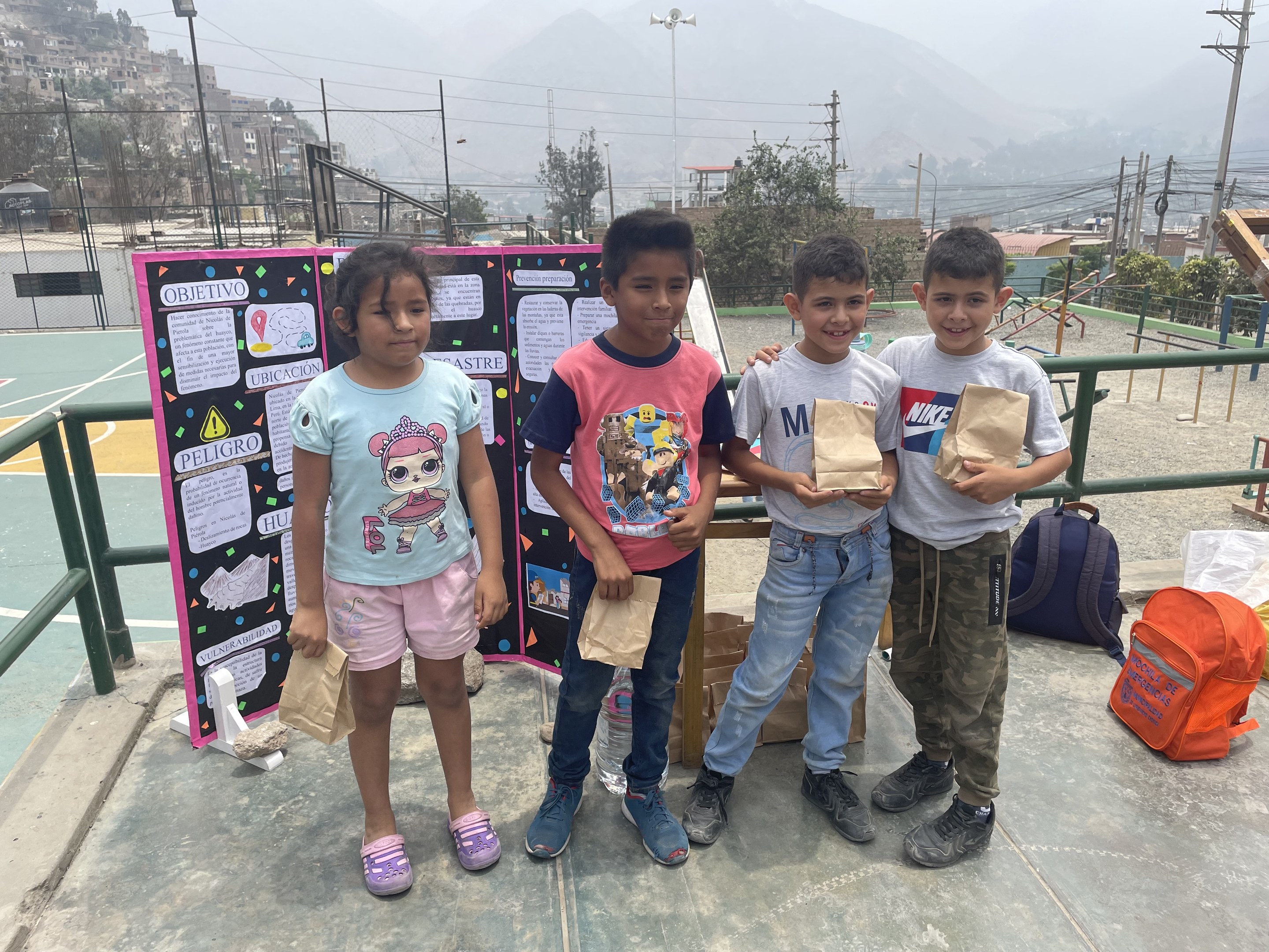 Gruppe von Kindern auf einem Basketballplatz mit Papiertüten in der Hand, mit Vorräten auf der rechten Seite und einer Tafel mit Text auf der linken Seite, vor einer Hintergrundkulisse aus Zäunen, Pfosten, Straßenlaternen, Bäumen, Gebäuden, Hügeln und Himmel.