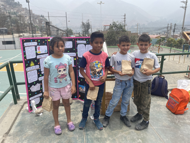 Gruppe von Kindern auf einem Basketballplatz mit Papiertüten in der Hand, mit Vorräten auf der rechten Seite und einer Tafel mit Text auf der linken Seite, vor einer Hintergrundkulisse aus Zäunen, Pfosten, Straßenlaternen, Bäumen, Gebäuden, Hügeln und Himmel.