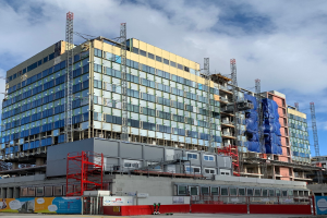 Großes Gebäude im Bau in einer städtischen Umgebung mit Glasfenstern, Metallrahmen, einer Treppe, einem Umzäunung, einem Schild, Strommasten, einer Straßenlaterne, nahen Bäumen und einem bewölkten Himmel.