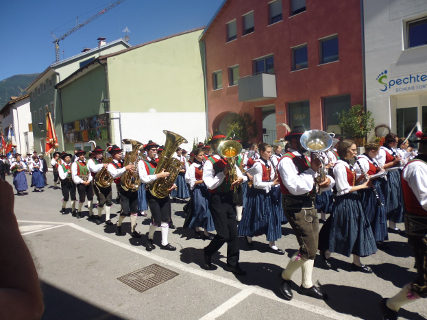 Eine Gruppe von Menschen in traditioneller bayrischer Tracht, die auf der Straße Musikinstrumente spielen, während sie an Gebäuden vorbeigehen, einige halten Fahnen, mit einem Hügel und einem klaren blauen Himmel im Hintergrund.
