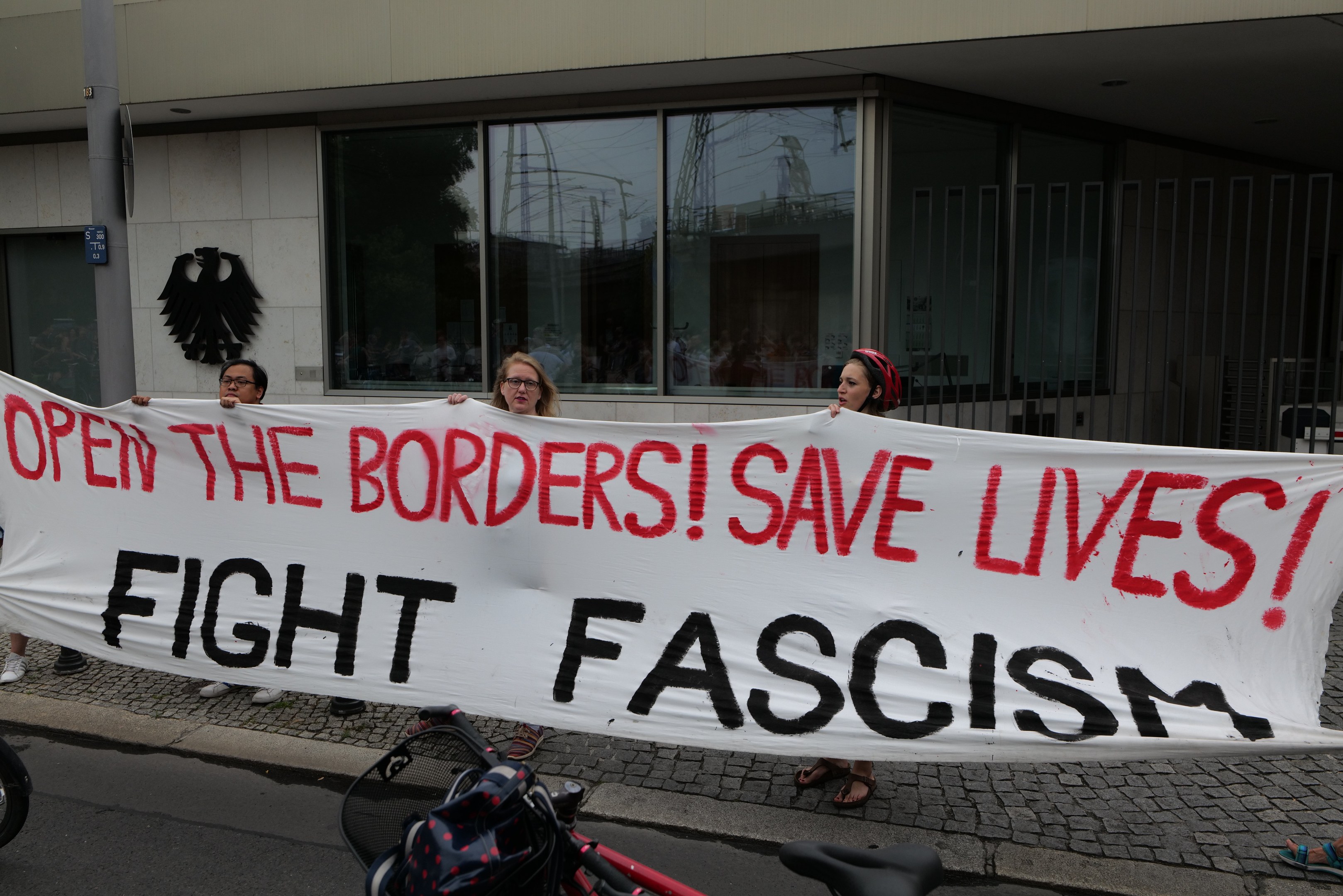 Menschen mit einem Transparent mit der Aufschrift "Öffnet die Grenzen, rettet Leben, kämpft gegen Faschismus" vor einem Gebäude mit Glasfenstern und einem Mast, mit einem Fahrrad und einer Tasche auf der Straße.