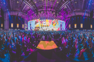 Eine große Menschenmenge sitzt auf Stühlen und schaut auf eine hell erleuchtete Bühne mit mehreren stehenden Personen, wahrscheinlich auf einem Online-Marketing-Rockstars-Event, umgeben von Bildschirmen.