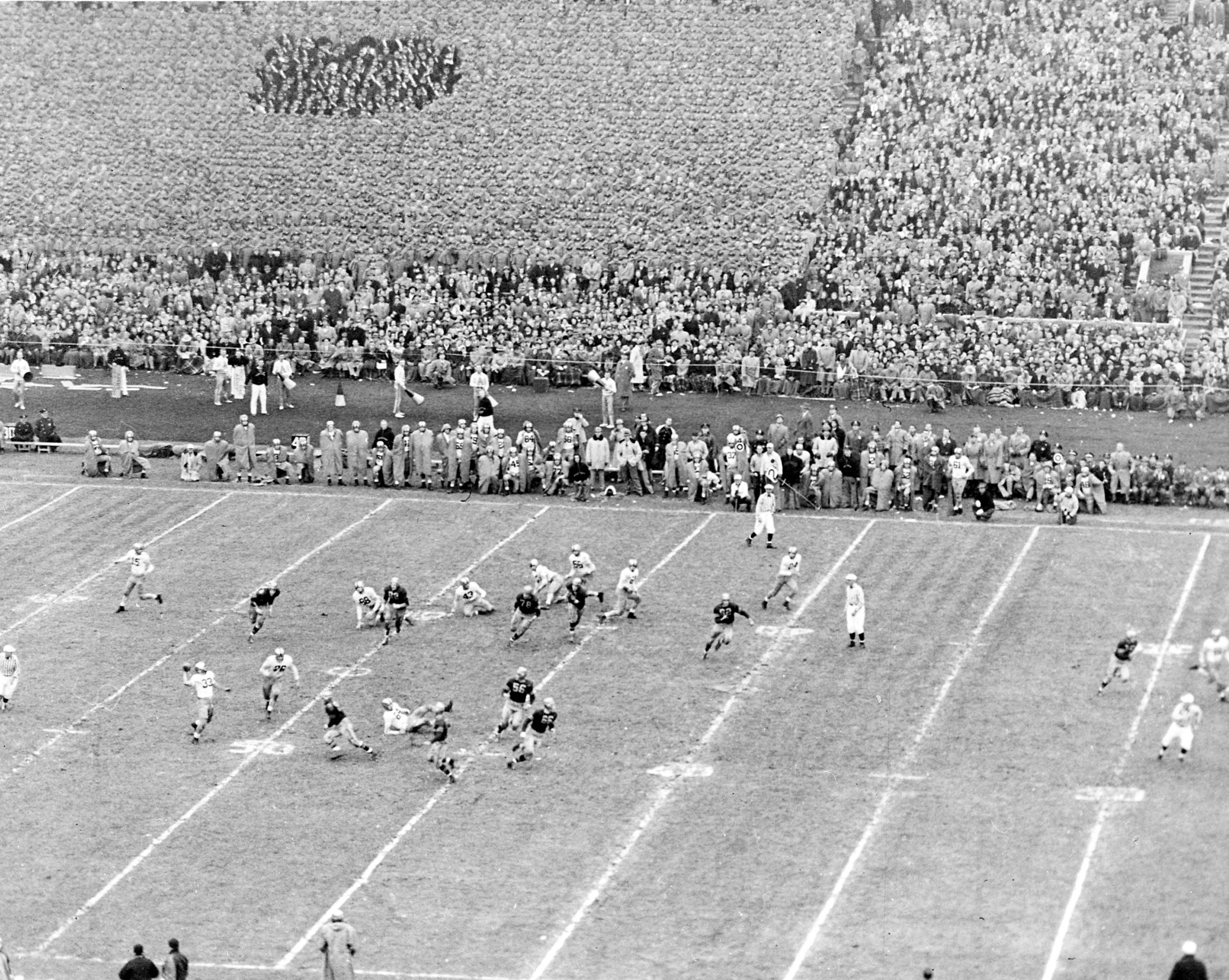 Schwarzes und weißes Foto eines Fußballspiels in einem Stadion mit Spielern auf dem Feld und Zuschauern in den Rängen.