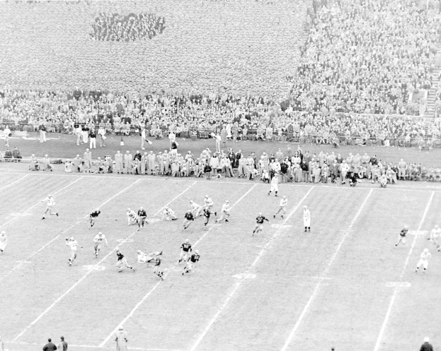 Schwarzes und weißes Foto eines Fußballspiels in einem Stadion mit Spielern auf dem Feld und Zuschauern in den Rängen.