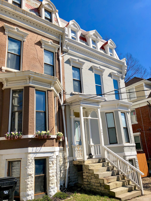 Eine Reihe von Stadthäusern mit Glasfenstern, Treppen mit Geländern, Blumentöpfen, Rasen, einem Müllcontainer, Stromleitungen, Bäumen und einem bewölkten Himmel.