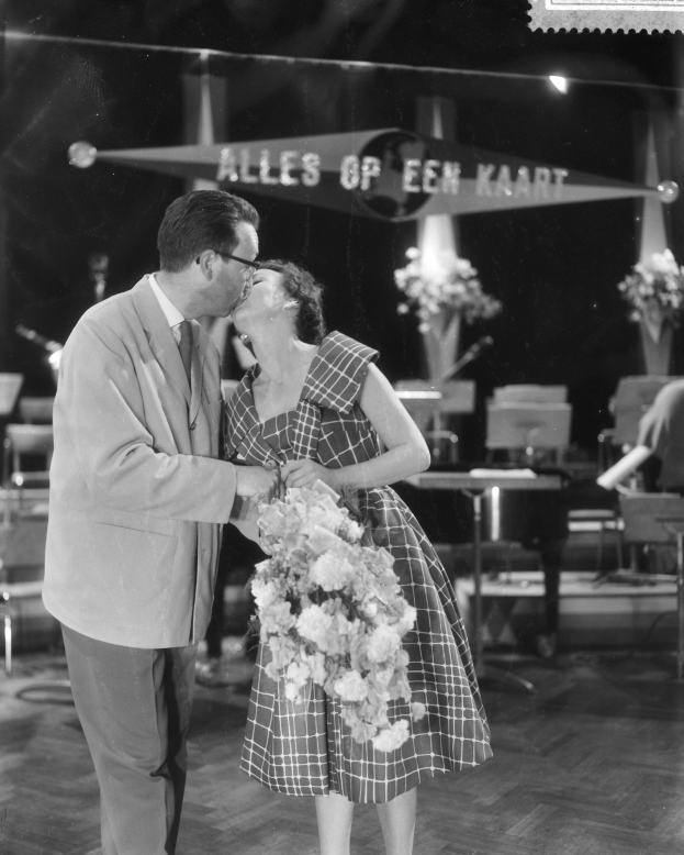 Ein Mann und eine Frau umarmen und küssen sich auf einer Bühne, wobei die Frau einen Blumenstrauß hält, während im Hintergrund Stühle und Blumenvasen zu sehen sind.