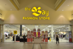 Ein Modegeschäft namens 'bay fashion' in einem Einkaufszentrum mit Einkäufern, Wandtafeln, Säulen, Deckenleuchten und Hintergrundauslagen.