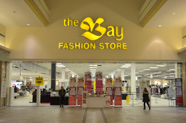 Ein Modegeschäft namens 'bay fashion' in einem Einkaufszentrum mit Einkäufern, Wandtafeln, Säulen, Deckenleuchten und Hintergrundauslagen.