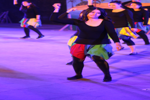 Eine Gruppe junger Frauen tanzt auf einer Bühne, mit einem Gemälde an der Wand im Hintergrund, alle tragen Schuhe.
