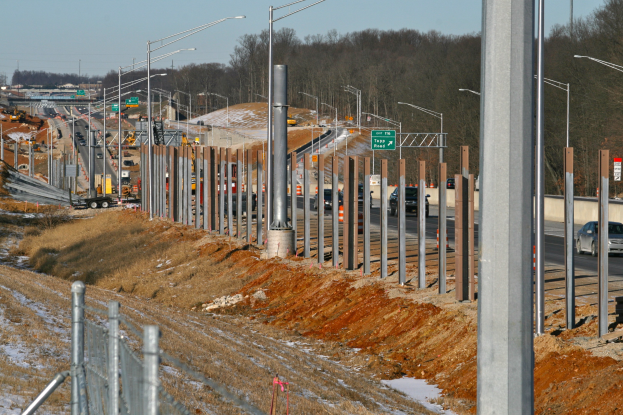 Baustelle mit mehreren Fahrzeugen, Pfählen, beleuchteten Schildern, einem Zaun, Gras- und Schneeflächen, Bäumen und einem Himmel im Hintergrund.