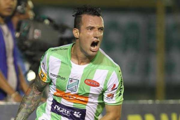 Ein Fußballspieler in grüner und weißer Uniform mit Tattoos auf dem Arm feiert ein Tor, während ein unscharfer Hintergrund Zuschauer und ein Schild zeigt.