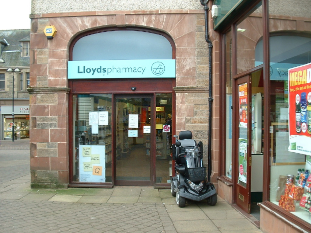 Eine Apothekenfiliale mit einem vor dem Gebäude geparkten Auto und einem Gebäude in der linken Ecke.