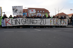 Eine Gruppe von Menschen hält ein Transparent mit der Aufschrift 'Kein Grund zu Feiern' und protestiert gegen deutsche Sparmaßnahmen auf einer Straße mit Gebäuden, Bäumen und einem klaren Himmel im Hintergrund.