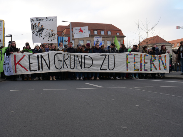 Eine Gruppe von Menschen hält ein Transparent mit der Aufschrift 'Kein Grund zu Feiern' und protestiert gegen deutsche Sparmaßnahmen auf einer Straße mit Gebäuden, Bäumen und einem klaren Himmel im Hintergrund.