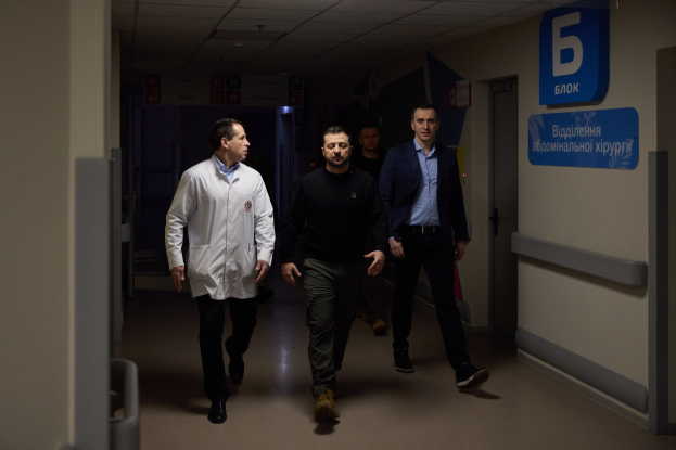 Eine Gruppe von Männern, die einen Krankenhausflur entlanggehen, mit Deckenleuchten, einer Tür an der rechten Wand und Texttafeln an der linken Wand, die besorgt aussehen.