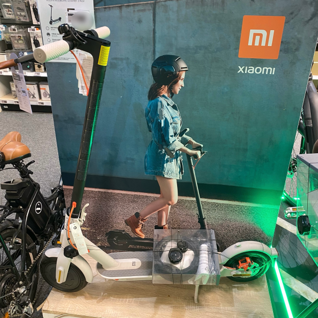 Eine Frau mit Helm fährt ein Xiaomi M365-Elektro-Scooter in einem Geschäft, rechts davon eine Glasvitrine mit verschiedenen Gegenständen und Anzeigetafeln im Hintergrund.