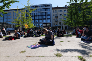 Eine Gruppe von Menschen sitzt vor einem Gebäude auf dem Boden während einer Demonstration in Berlin, einige tragen Masken, mit verstreuten Taschen und Gegenständen, unter einem klaren blauen Himmel.