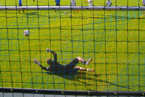 Ein Fussballspieler taucht ab, um einen Ball vor dem Tor zu halten, mit einem Netz im Vordergrund und einigen Menschen im Hintergrund, während mehrere Fussbälle auf dem Boden verstreut sind.