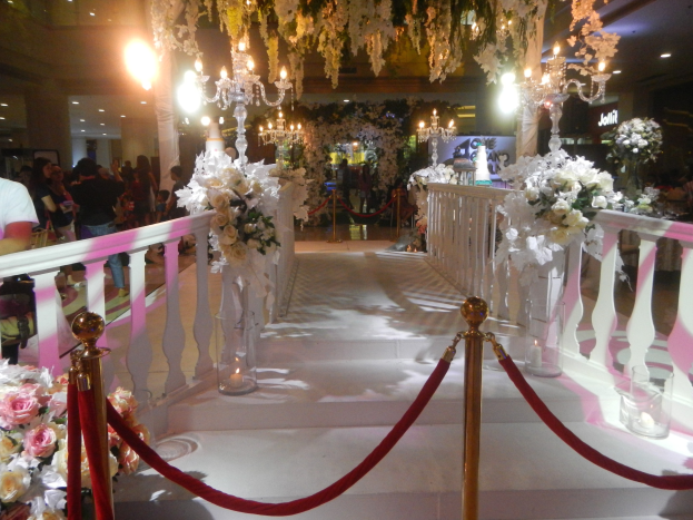 Eine Trauung in einem Einkaufszentrum mit weißem Teppich, Dekorationselementen, Blumenarrangements und Deckenbeleuchtung, bei der zahlreiche Gäste anwesend sind.