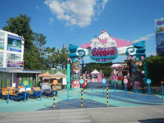Eine Karussellfahrt am Eingang eines Parks mit einer Straße, einem Zaun, Buden, Fahnen, Bäumen und einem bewölkten Himmel im Hintergrund.