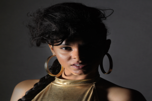 Eine Frau in einem goldenen Kleid und großen Kreolen steht selbstbewusst vor einer Wand, ihre glatten Bob-Haare sind ordentlich frisiert, sie trägt dezenten natürlichen Make-up mit pinkem Rouge und glänzenden Lippen und schaut direkt in die Kamera mit einem starken, entschlossenen Ausdruck.