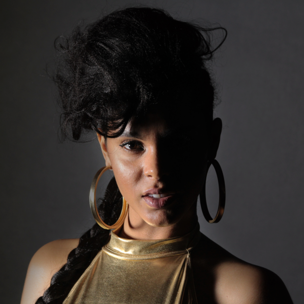 Eine Frau in einem goldenen Kleid und großen Kreolen steht selbstbewusst vor einer Wand, ihre glatten Bob-Haare sind ordentlich frisiert, sie trägt dezenten natürlichen Make-up mit pinkem Rouge und glänzenden Lippen und schaut direkt in die Kamera mit einem starken, entschlossenen Ausdruck.