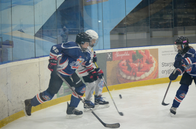 Gruppe junger Menschen, die Eis hockey auf einer Indoor-Eisfläche spielen, tragen Helme, Sportuniformen und Schlittschuhe und halten Hockey-Schläger, mit einer Glaswand und einem Plakat im Hintergrund.