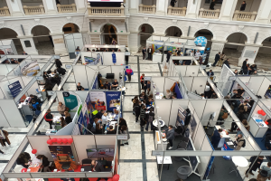Große Menschenmenge auf einer internationalen Fachmesse mit Ständen, Bannern, Tischen, Stühlen, Ballons und einem Gebäude mit Bögen, Säulen, Geländern und einem Bildschirm im Hintergrund.