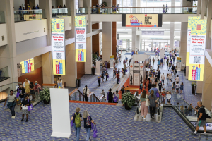 Eine große Menschenmenge bewegt sich durch ein großes Gebäude mit Bannern, Topfpflanzen, Rolltreppen, Geländern, Säulen, bedruckten Tafeln, Glastüren und anderen Elementen, einige tragen Taschen, was auf den Besuch der Nationalen Buchmesse hindeutet.